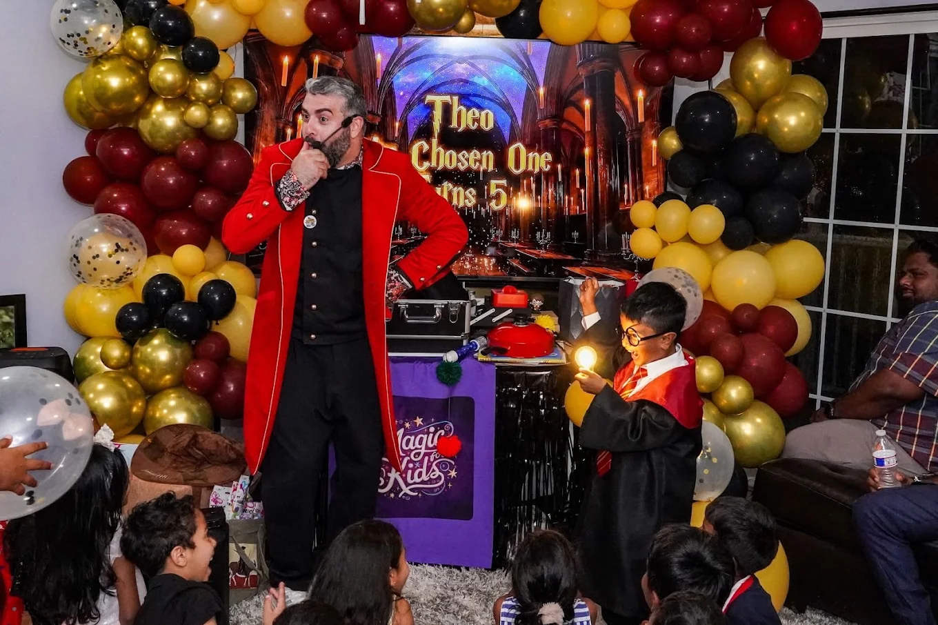 Nico in a red magician’s coat performing with a child as children watch at an indoor birthday party.